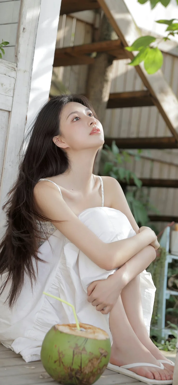 一名身穿白裙的女子坐在户外木平台上，身旁放着一只椰子。