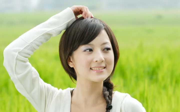 一名短发女性站在绿色田野前，抬手触碰头发并面带微笑。