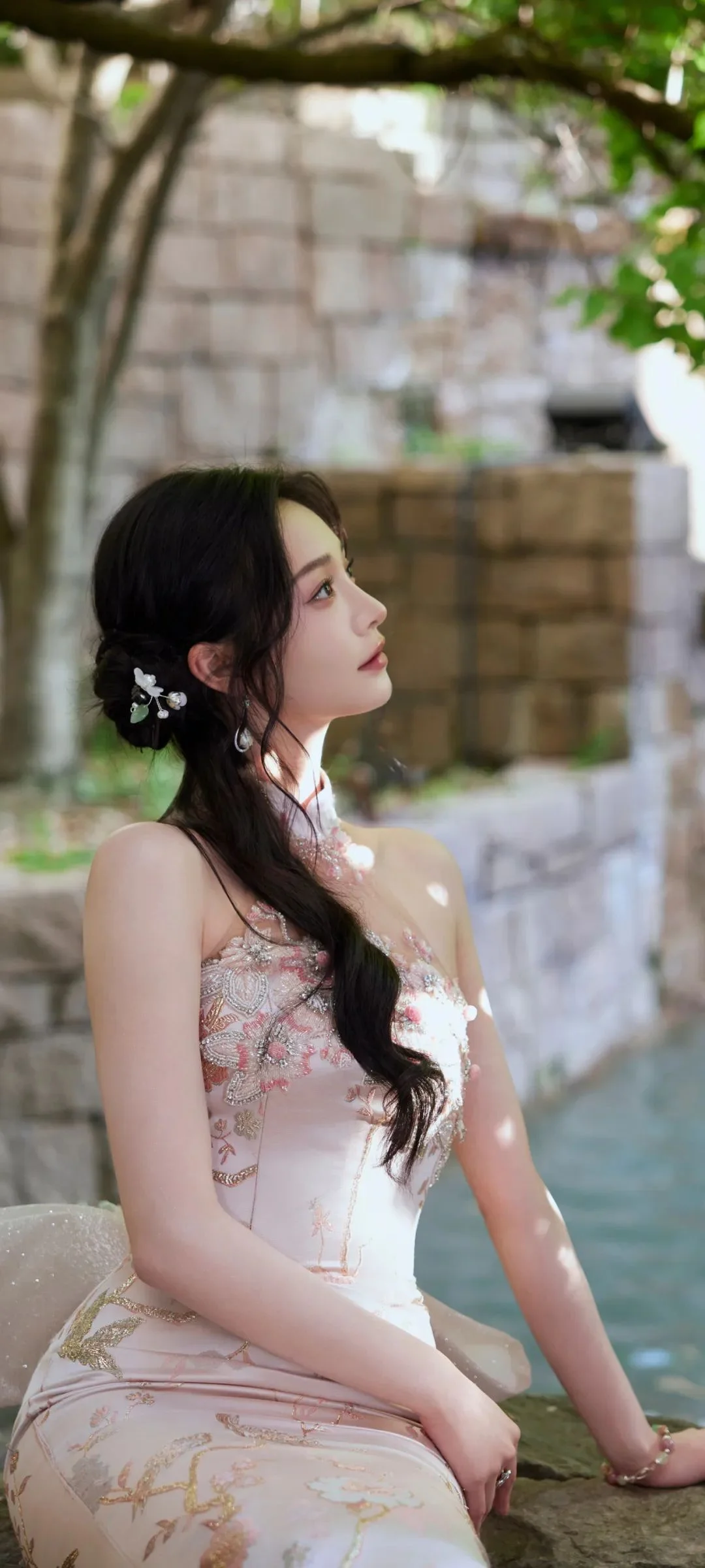 長い髪の女性が花柄の淡いドレスを着て、木陰の水辺に屋外で座っています。