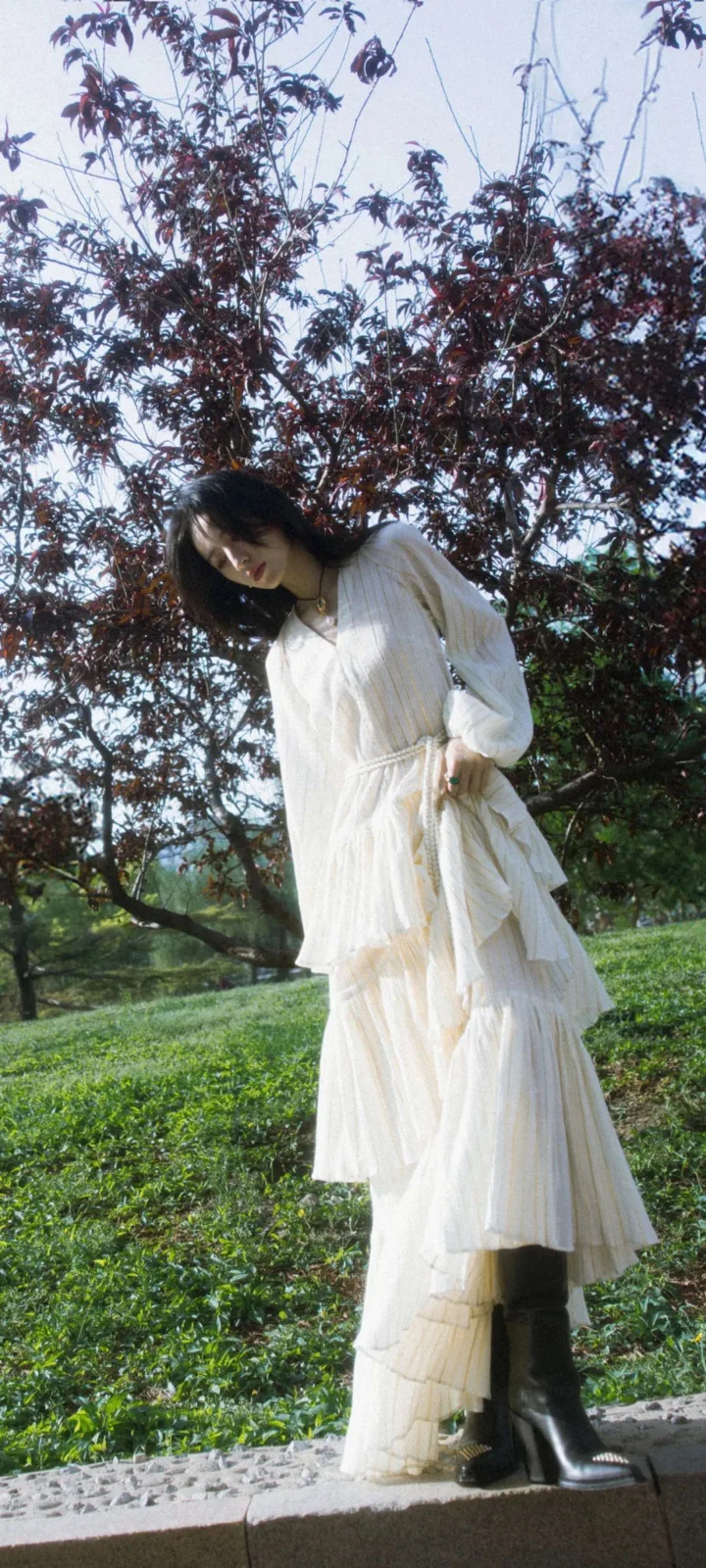 흰색 긴 드레스를 입은 여성이 나무와 잔디 옆에 서 있다.