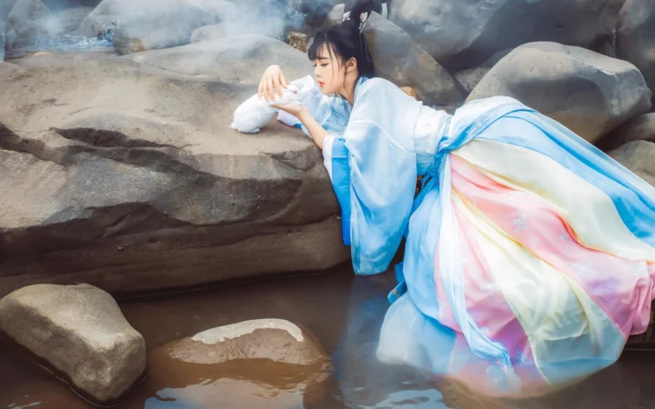 一名身穿浅蓝色传统服饰的女子倚坐在溪水与岩石之间。