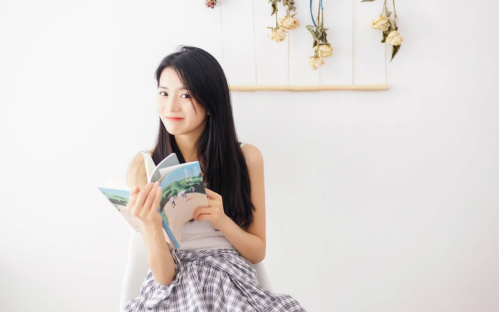 明るい室内で女性が開いた本を手に持ち、カメラに微笑みかけています。