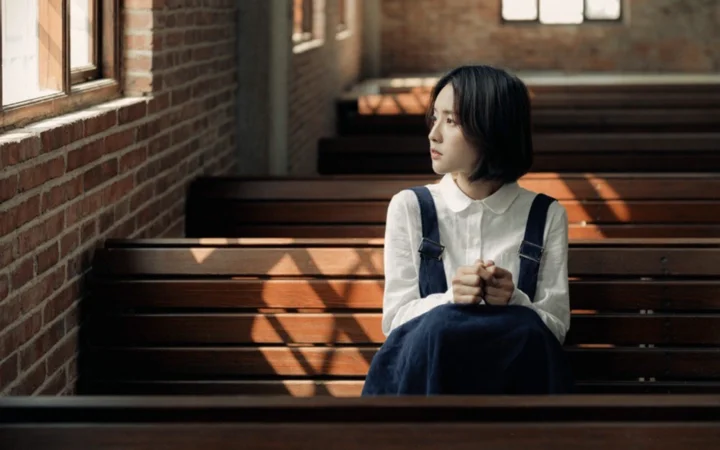 一名短发女子坐在砖墙窗边的木长椅上，阳光在室内投下斑驳光影。