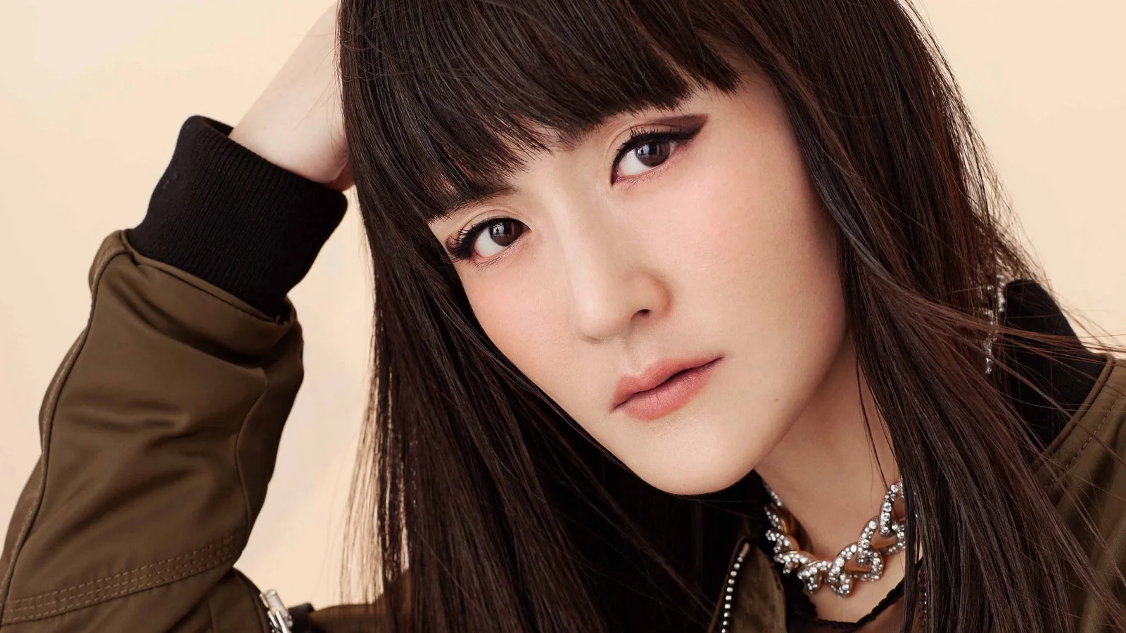 暗い色の長い髪の女性が、茶色のジャケットを着てカメラに向かうクローズアップポートレート。