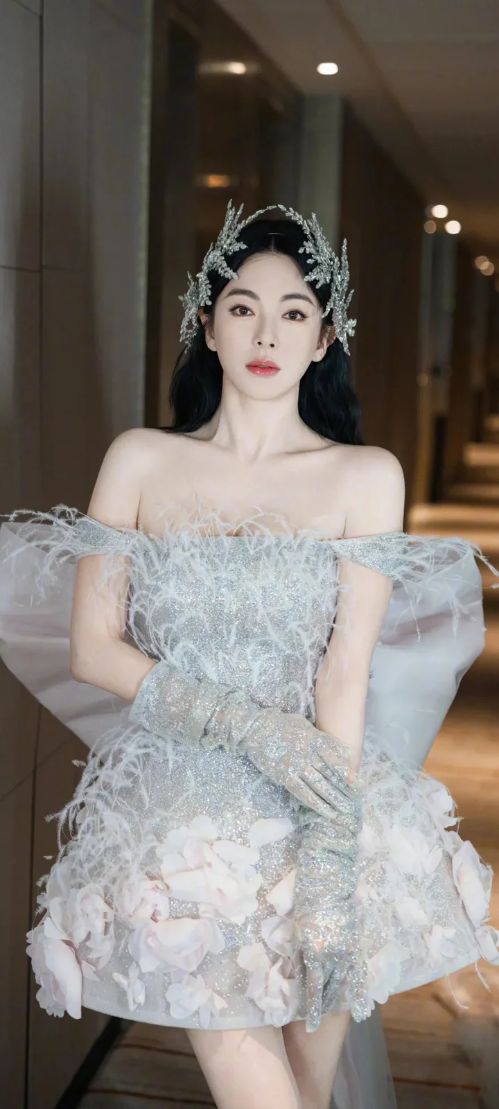 一位成年女性身穿銀白色羽飾禮服站在室內走廊中。