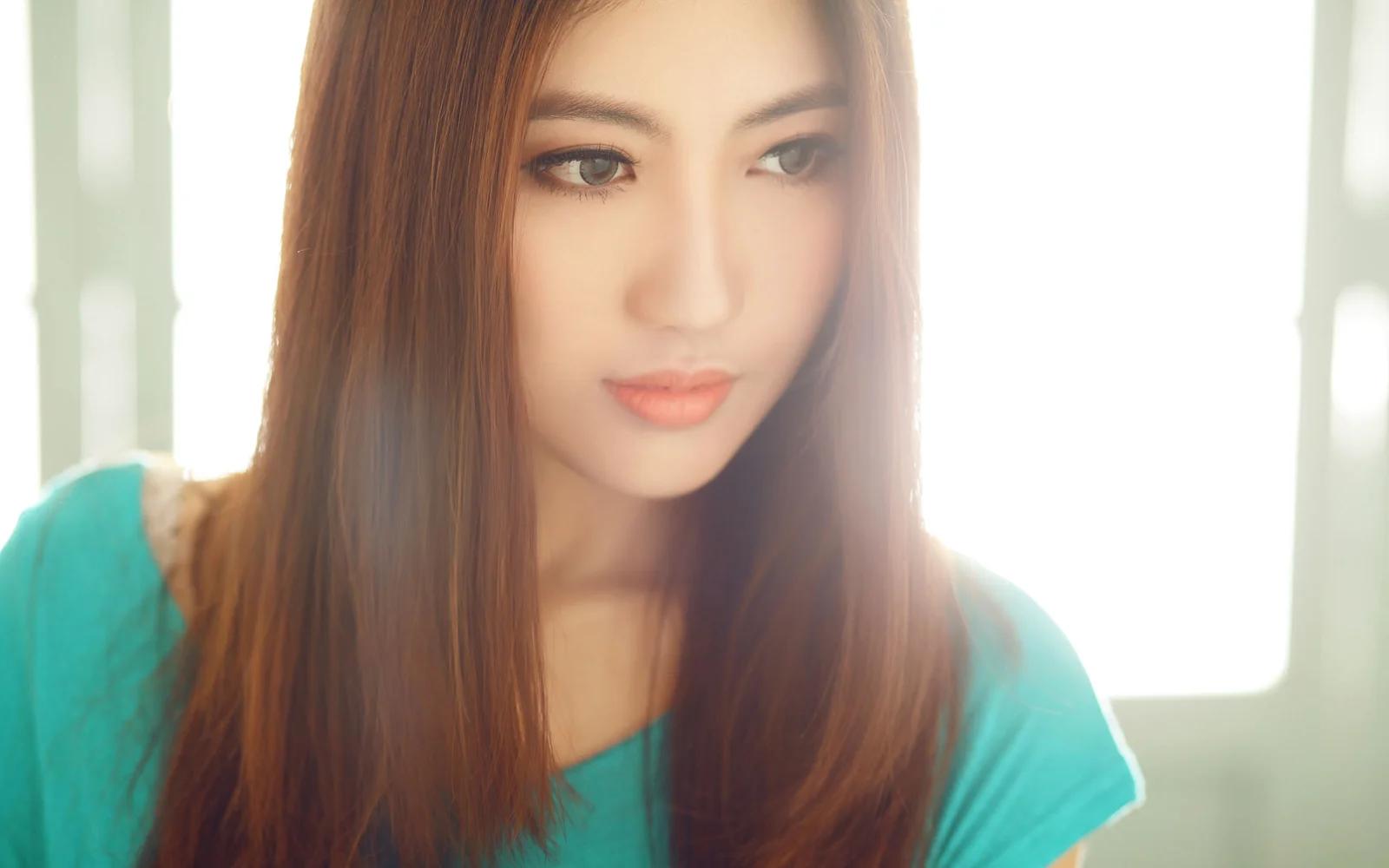긴 머리의 여성이 청록색 상의를 입고 밝은 창가에 서 있습니다.