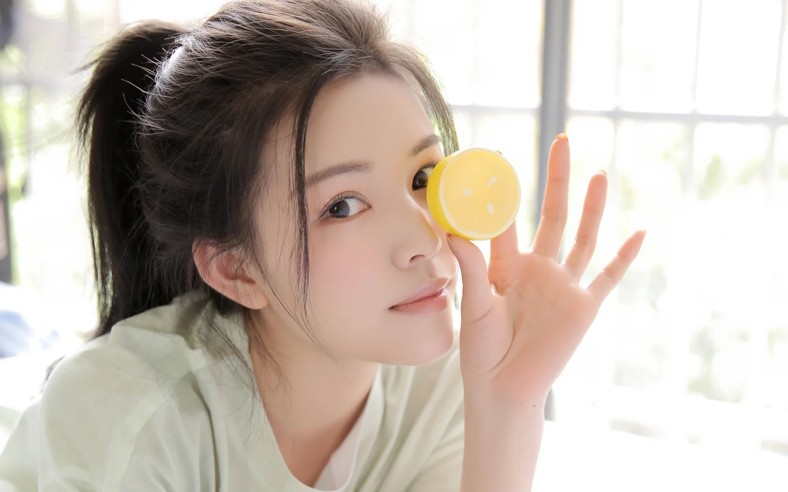 밝은 창가에서 여성이 노란 레몬을 들고 카메라를 향해 미소 짓고 있다.