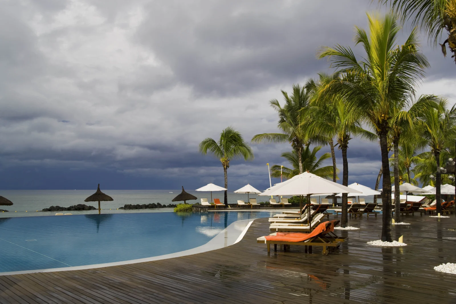 棕榈树环绕的海滨泳池旁设有遮阳伞和躺椅，远处海面上空乌云密布。