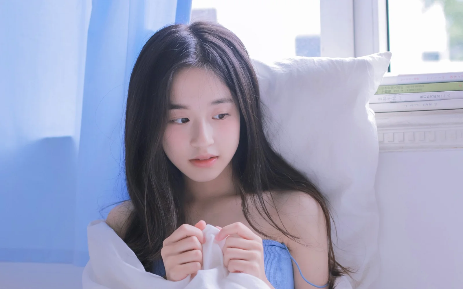 一名长发女性坐在窗边的床上，手持白色布料，画面以柔和的蓝白色调为主。