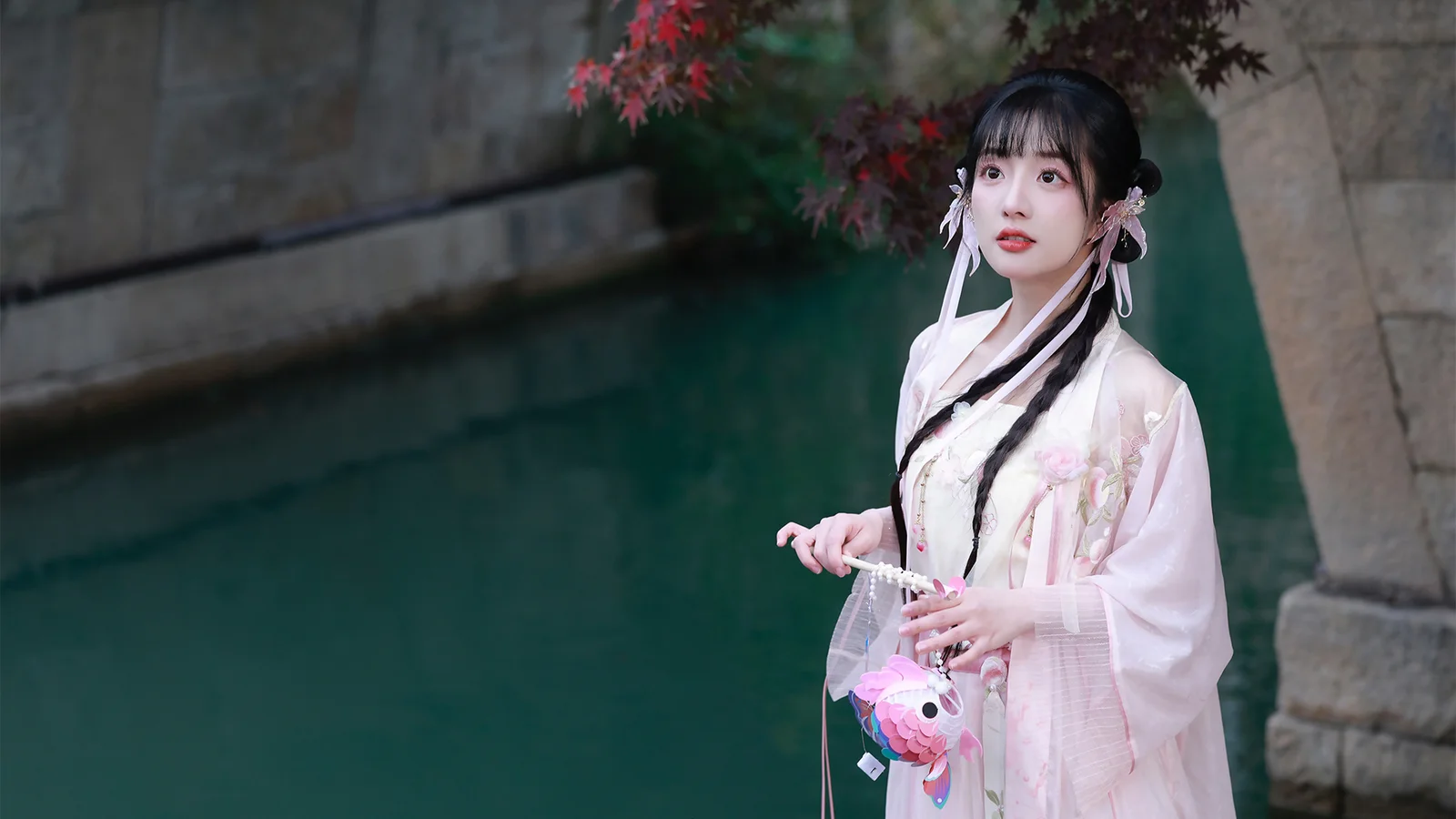 연분홍 한푸를 입은 여성이 초록빛 물과 돌담 옆에 서 있습니다.