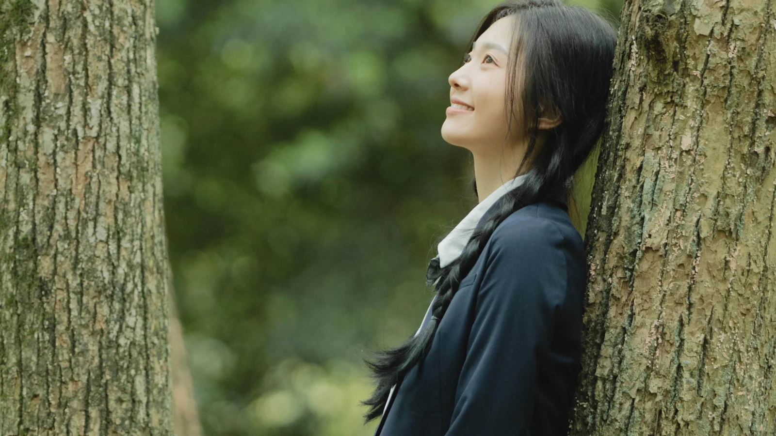 森の中で女性が木の幹にもたれ、見上げながら微笑んでいる。