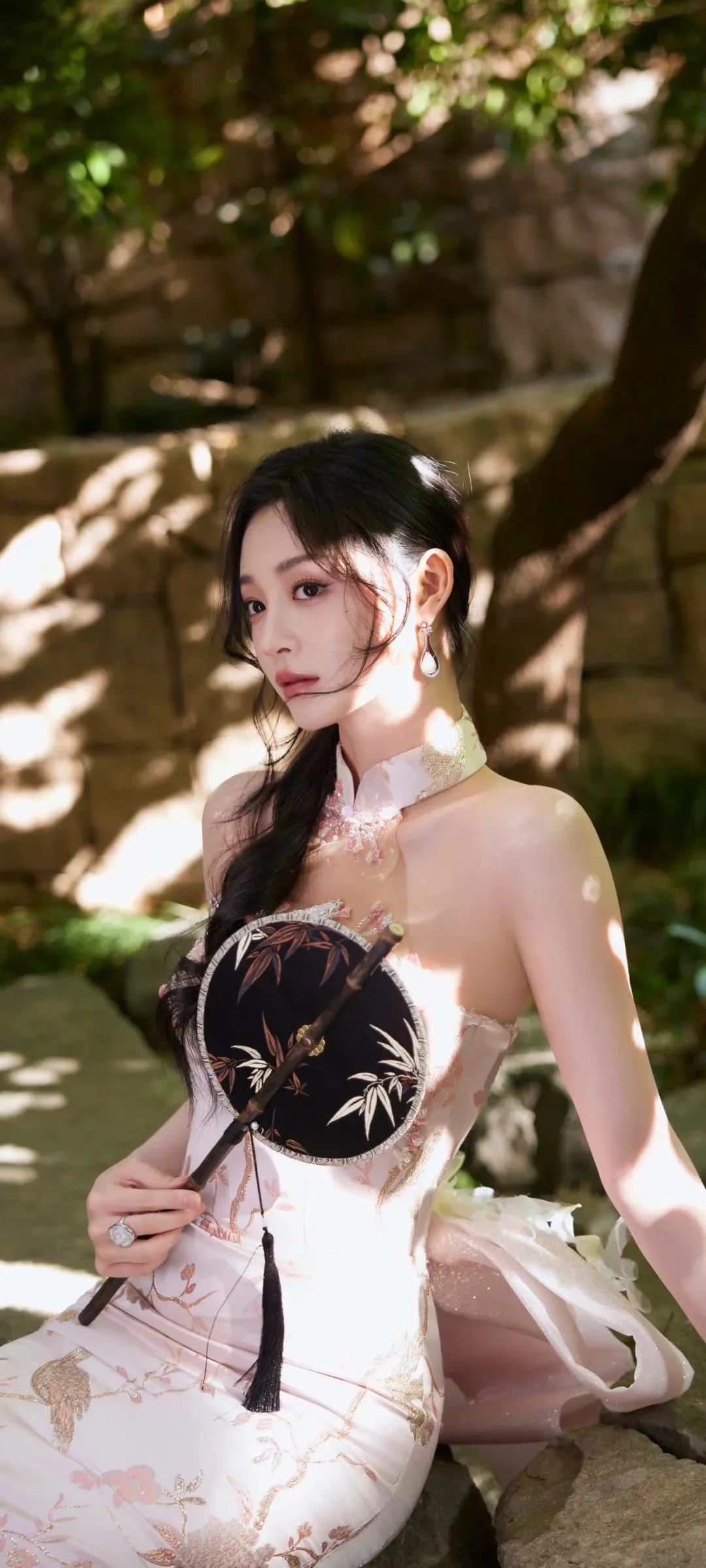 전통풍 의상을 입은 여성이 나무 그늘과 돌담 옆에 앉아 둥근 부채를 들고 있다.