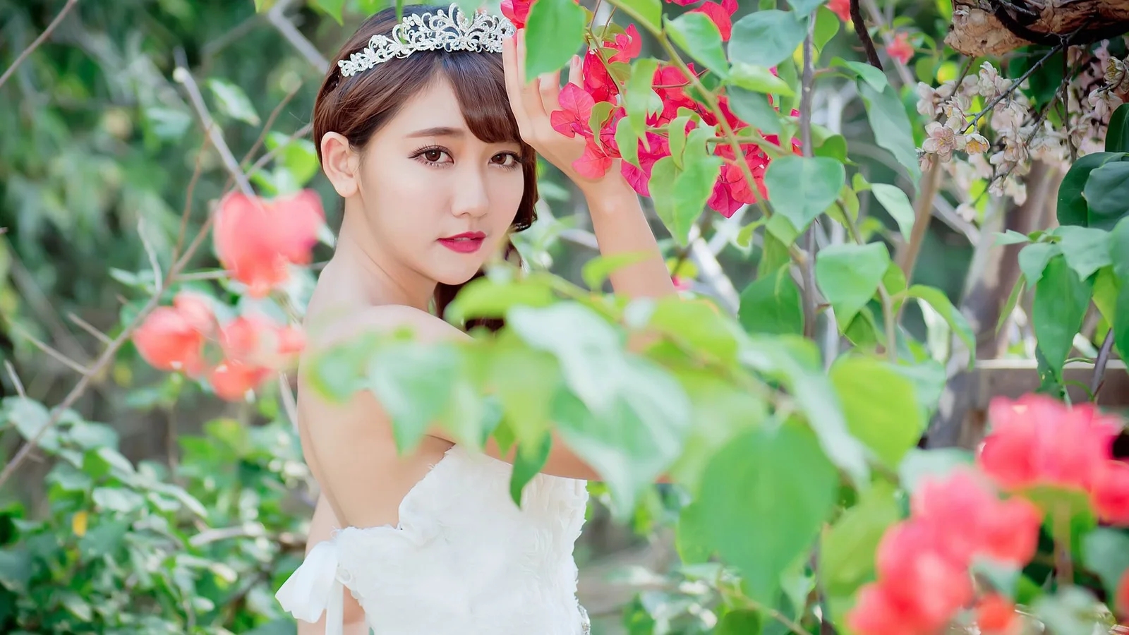 一名戴着头饰的女子穿着白色礼服站在繁花绿叶之间。