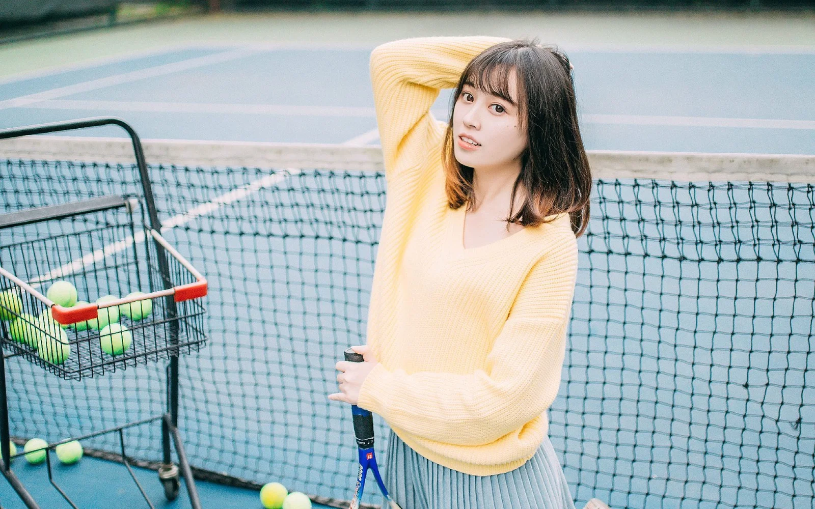 一名穿黄色针织衫和百褶裙的成年女性站在网球场旁，手持网球拍，旁边放着装有网球的球车。