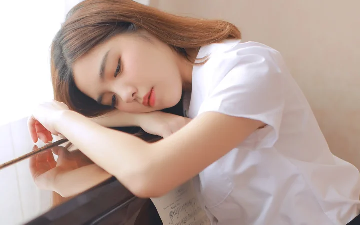 흰색 상의를 입은 여성이 피아노에 옆으로 기대어 차분하고 부드러운 표정을 짓고 있다.
