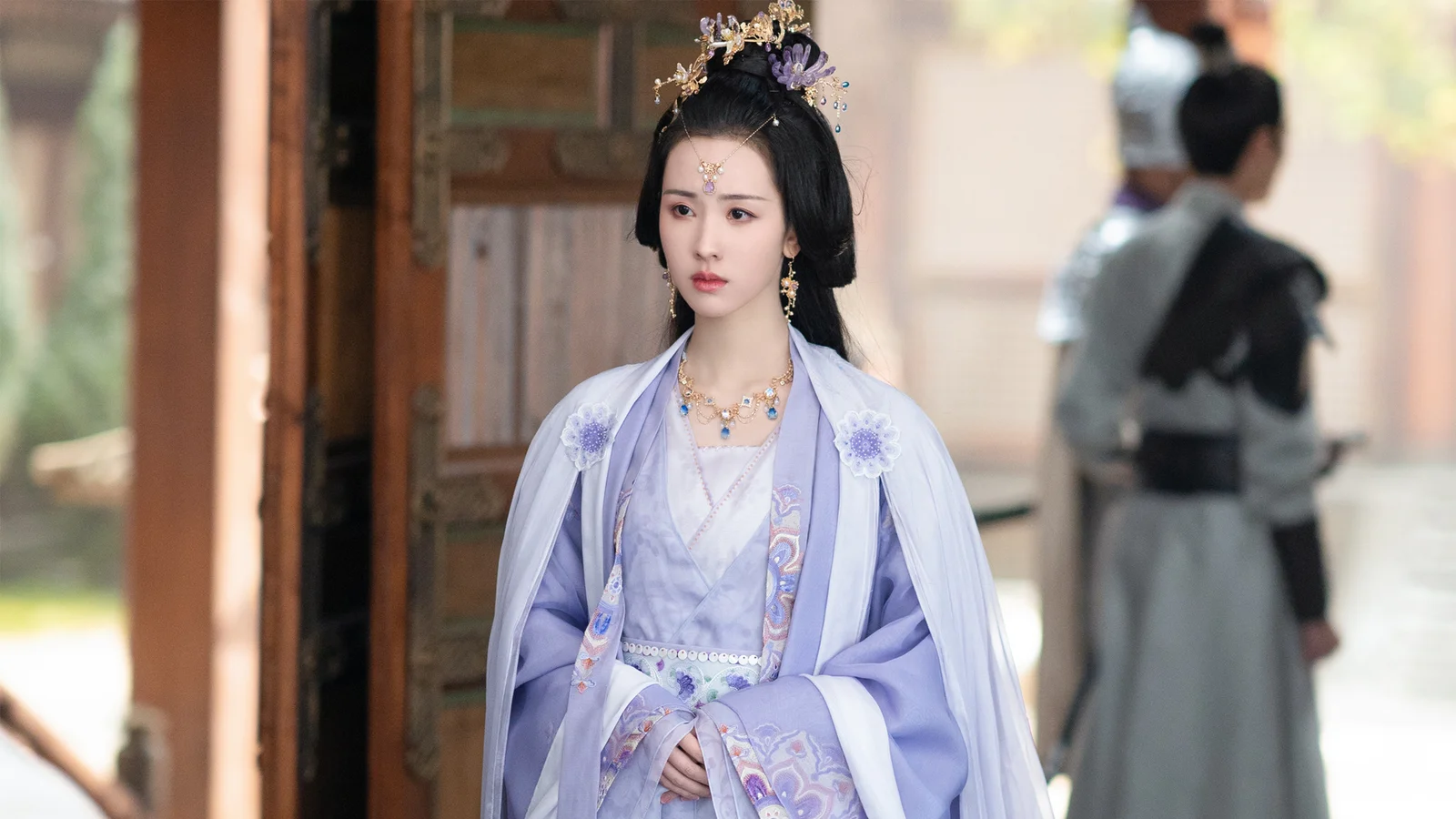 一名身穿淡紫色传统服饰、佩戴华丽头饰的女子站在木门旁。