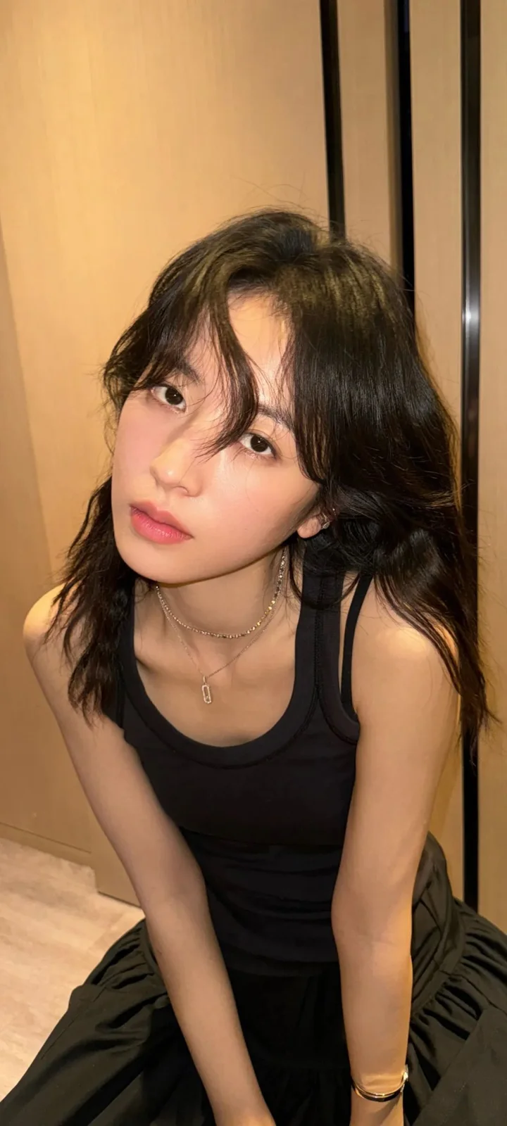 一位长发女性穿着黑色背心坐在室内木质背景前。