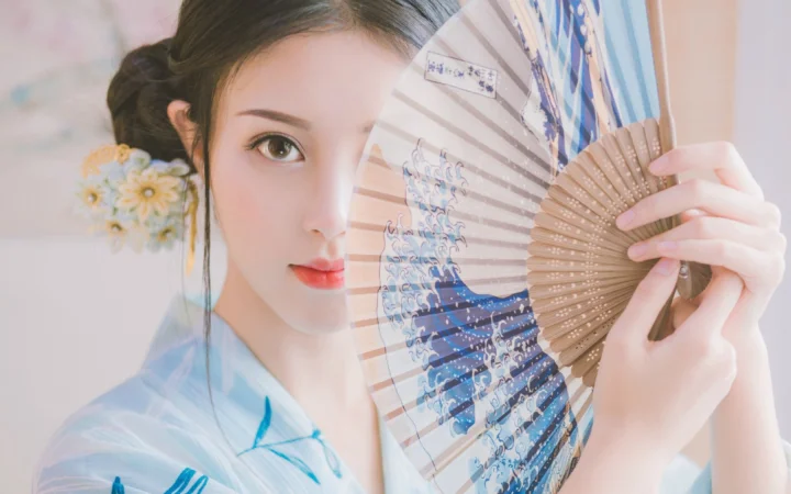 一位成年女子手持折扇面对镜头，画面呈现柔和的古风人像氛围。