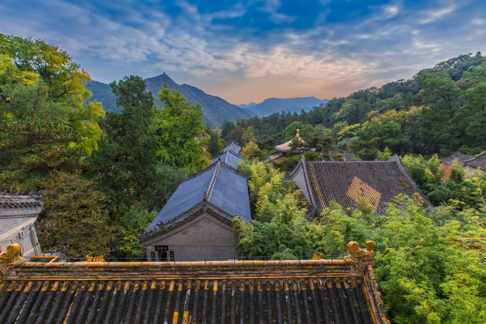 夕阳映照下，群山、茂密森林与传统建筑屋顶构成宁静的风景画面。
