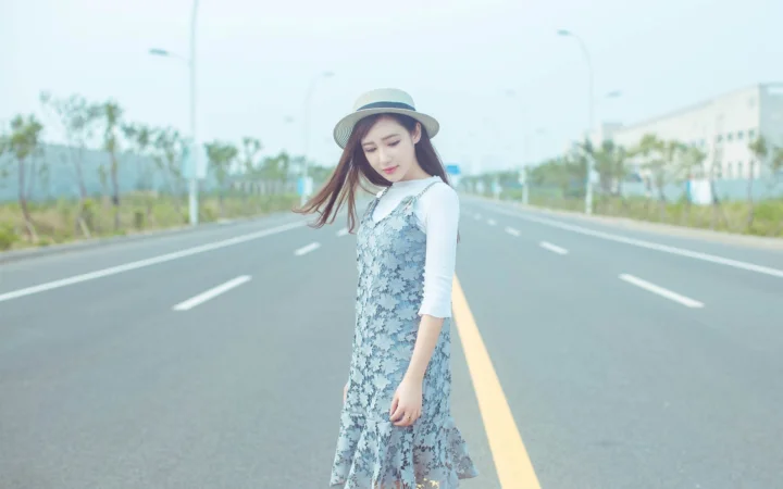 一名戴着帽子的女子站在宽阔道路中央，背景是淡蓝色天空和道路两侧的树木与建筑。