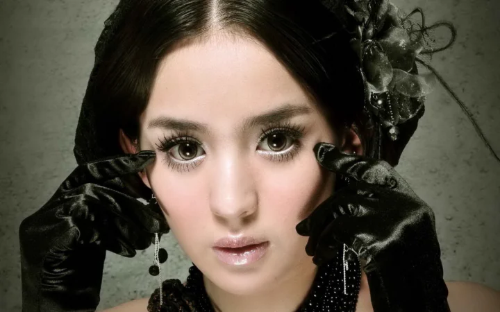 黒い手袋と暗色のヘッドピースを身に着けた女性を、灰色の背景の前で近距離から捉えたポートレートです。