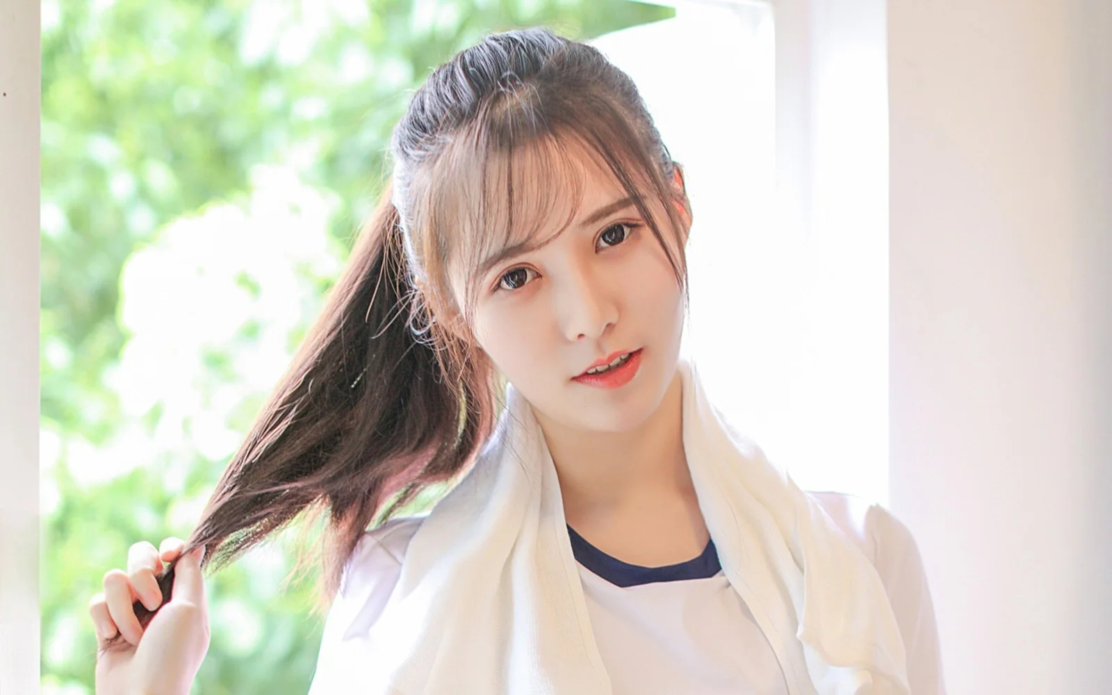 明るい窓辺でポニーテールの女性が髪先に触れながらカメラを見つめている。