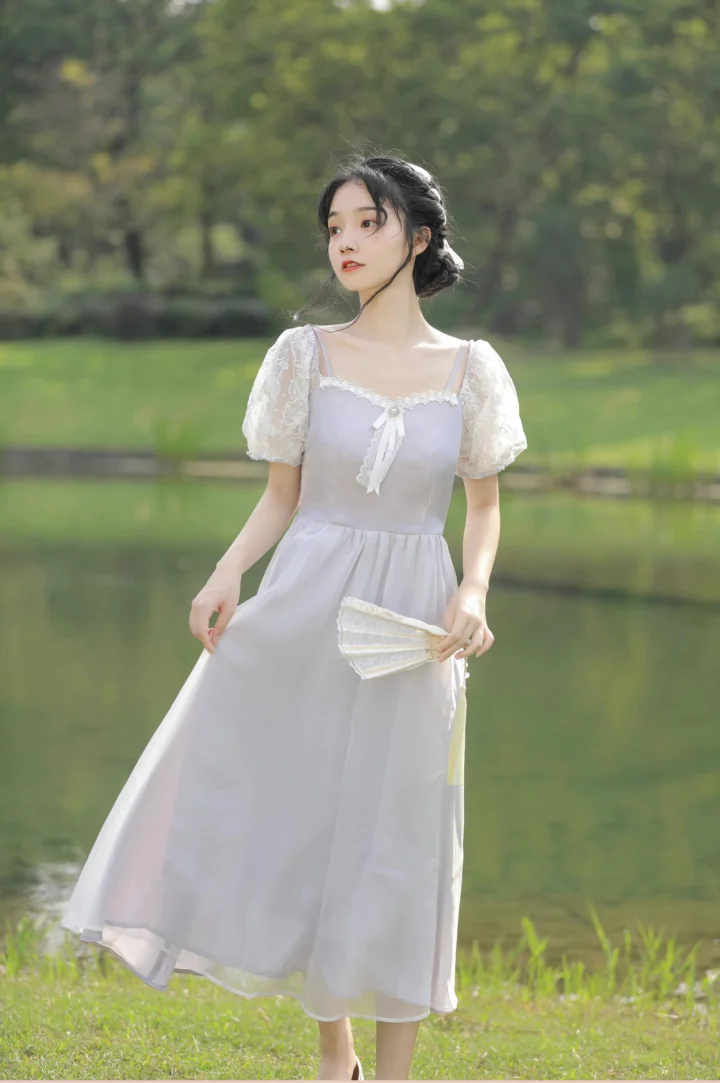 一名身穿白色连衣裙的女子手持折扇站在池塘边的草地上。