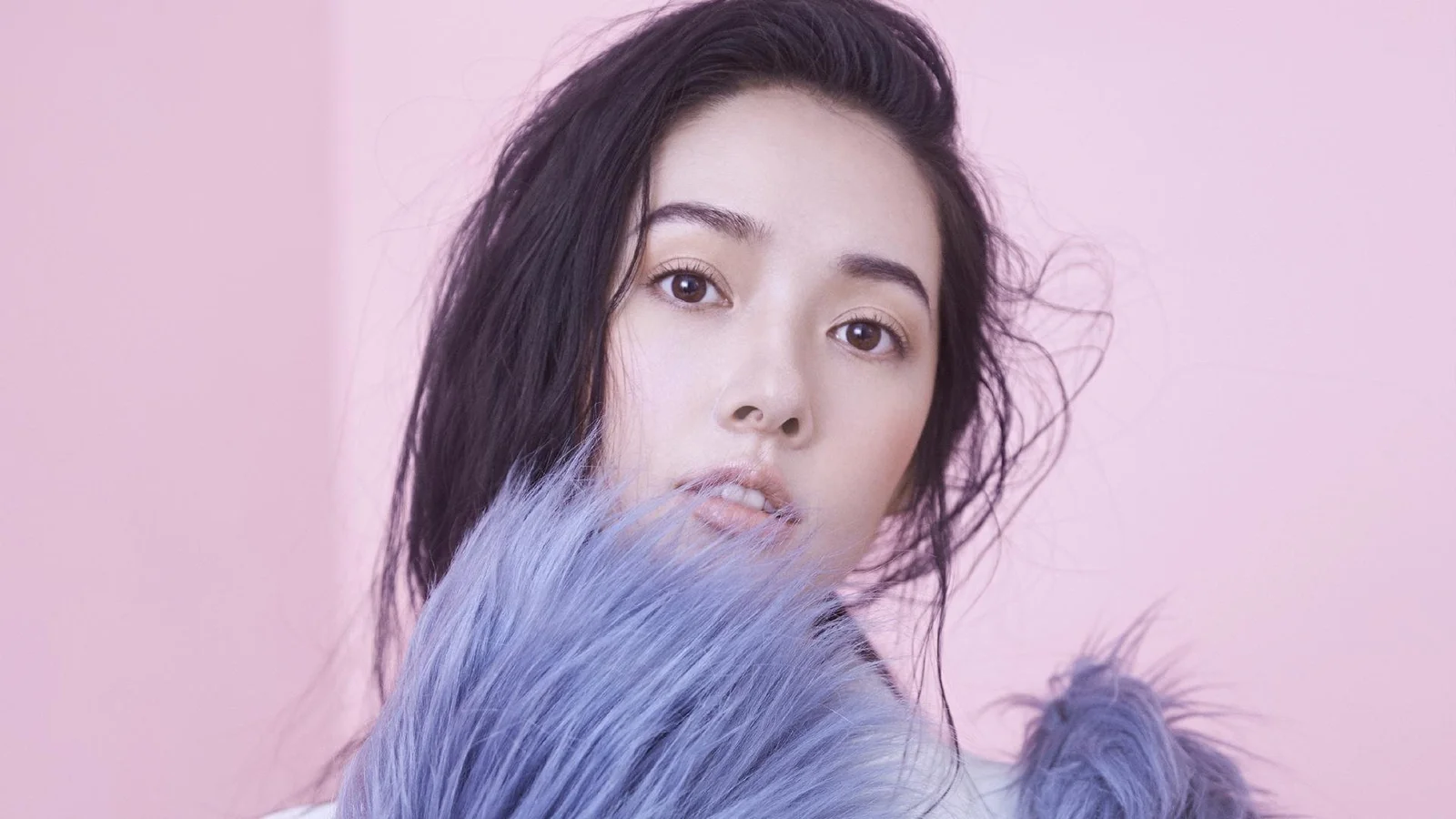 一名成年女性在粉紫色背景前被柔和光线照亮，前景环绕着紫色毛绒材质。