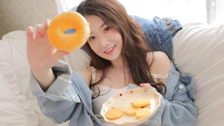 미소 짓는 성인 여성이 흰 침대 위에서 오렌지를 카메라 쪽으로 들고 있으며 앞에는 쿠키 접시가 놓여 있습니다.