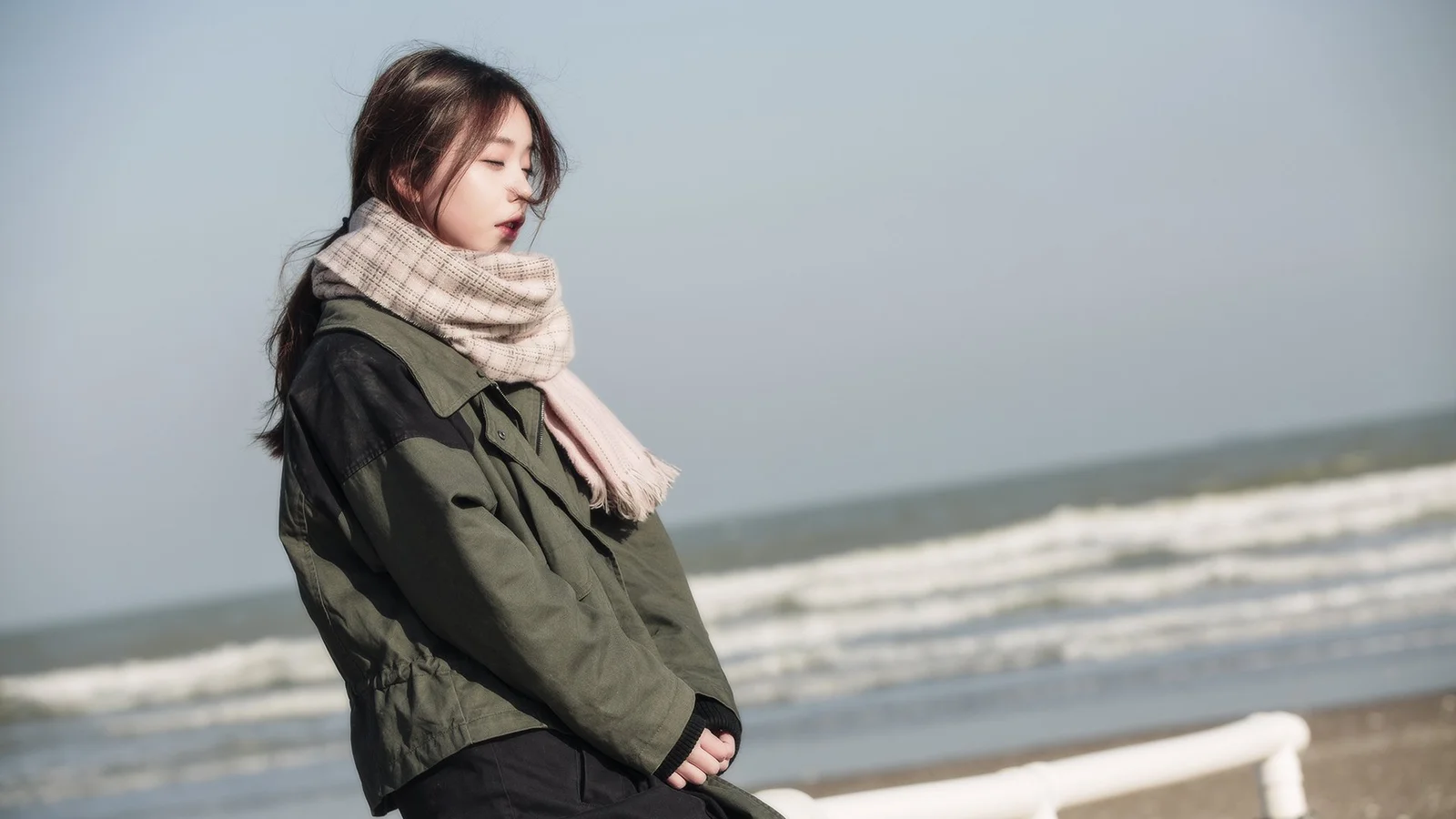 스카프를 두른 여성이 잔잔한 바다와 하얀 파도를 뒤로하고 해변에 서 있습니다.