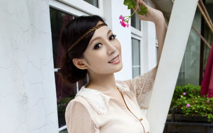 一名成年女子站在窗边，手持一枝粉色小花微笑面对镜头。