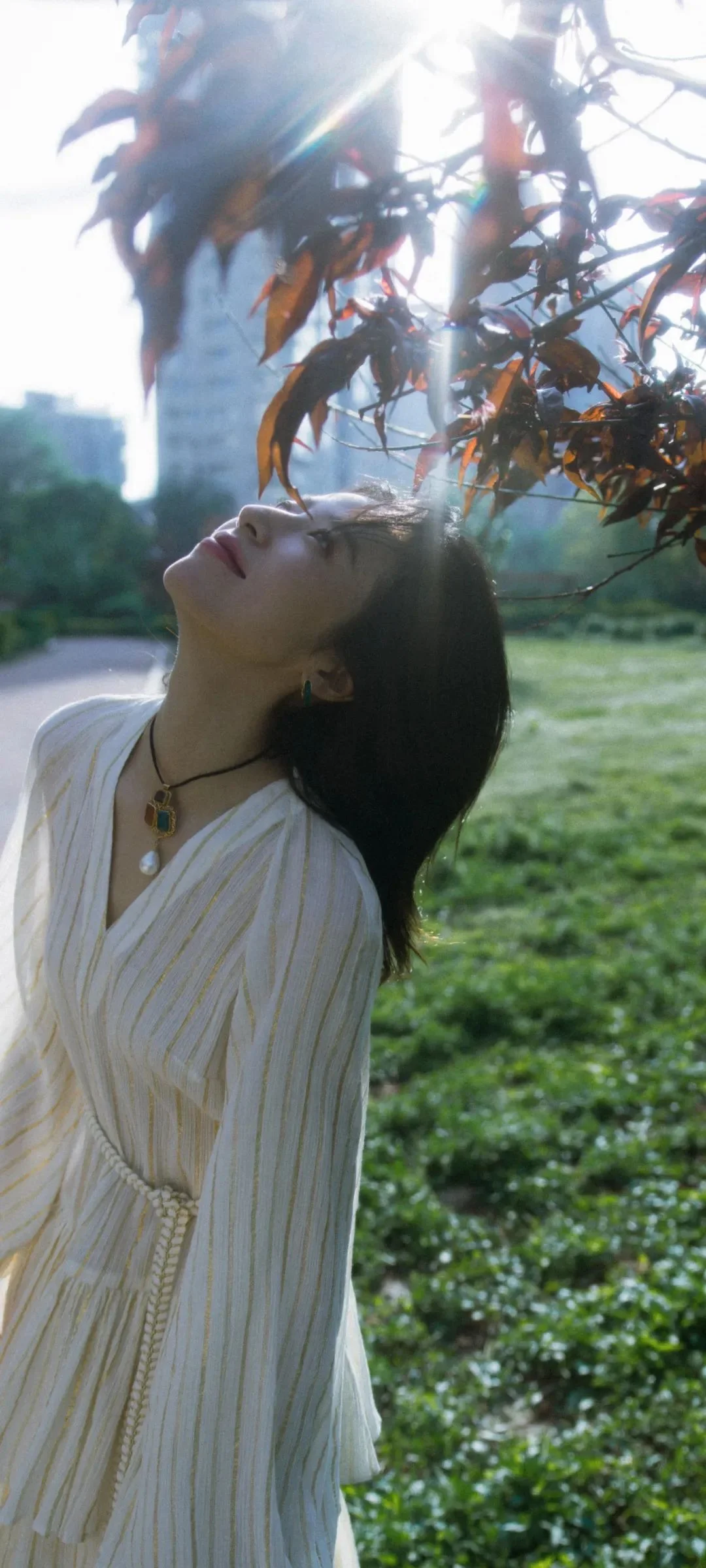 一名身穿白色长裙的女性在树枝和阳光下抬头站立，背景是草地与模糊的建筑。