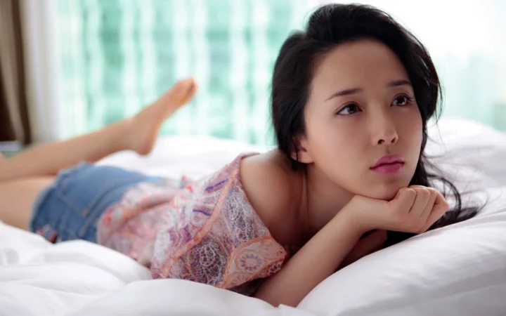 一名女性侧卧在白色床上，双手托着下巴望向一侧。