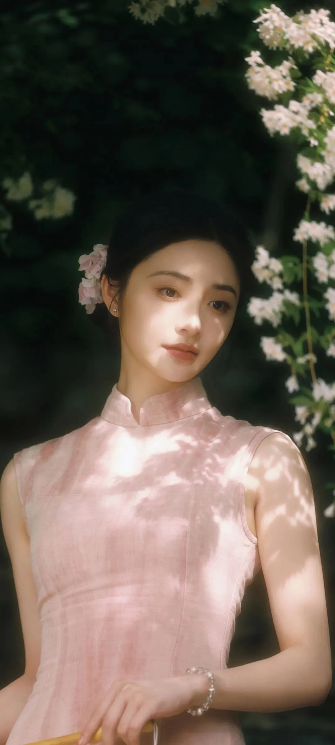 一名身穿粉色無袖裙裝的女子站在花叢與綠葉之間，臉上灑著斑駁的陽光。