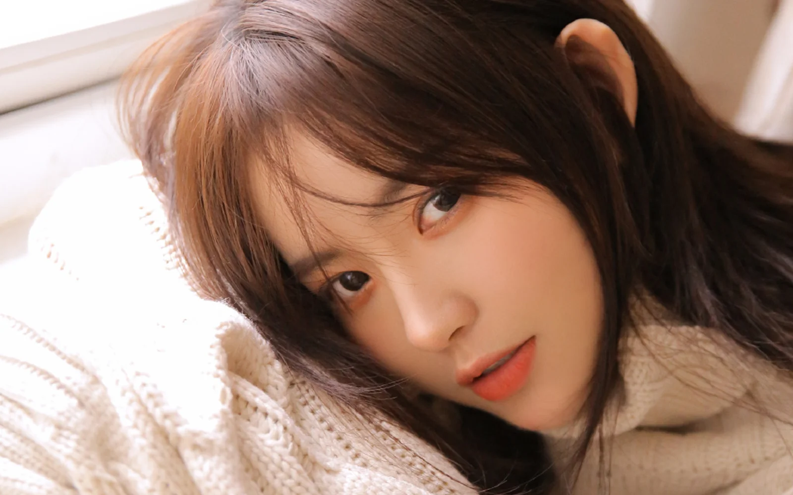 크림색 니트를 입은 갈색 머리 여성이 가까이에서 카메라를 바라보고 있다.