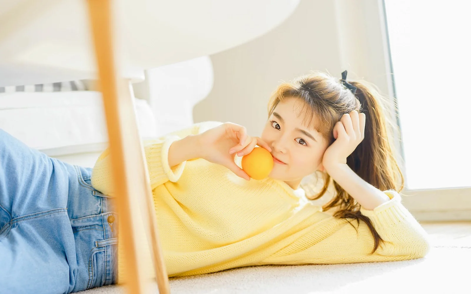 黄色いトップスの女性が、明るい室内でオレンジを手にして横たわっています。