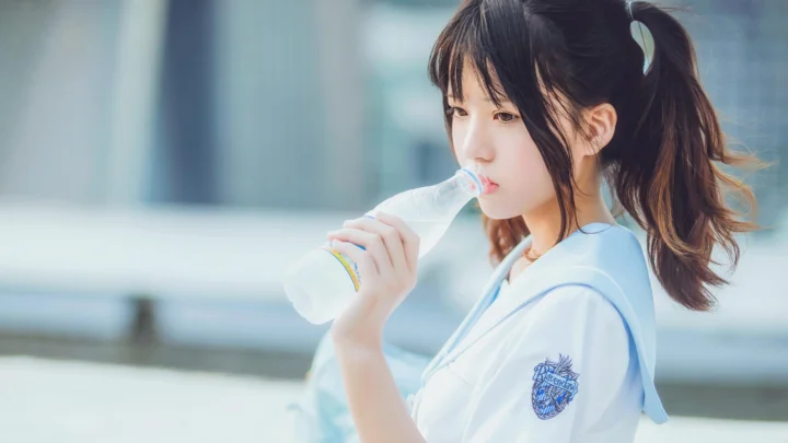 一名扎着马尾的年轻女性在户外拿着水瓶饮水。