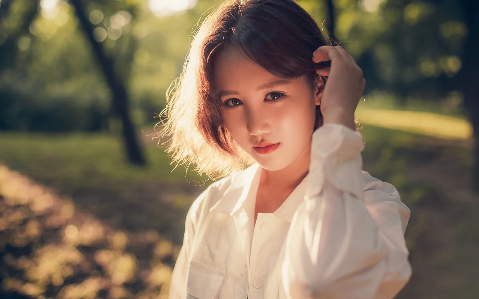 一位成年女性身穿白色上衣站在阳光斑驳的公园中。