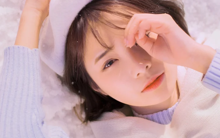 연보라색 니트를 입은 여성이 하얗고 푹신한 표면에 누워 얼굴 가까이에 팔을 둔 채 카메라를 바라보고 있다.