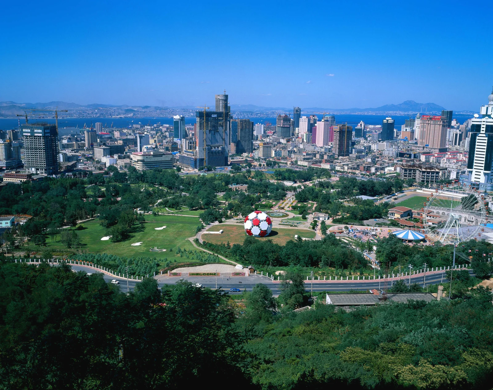 宽阔的城市公园位于前景，远处是密集的高层建筑和蓝色天空，右侧可见摩天轮。