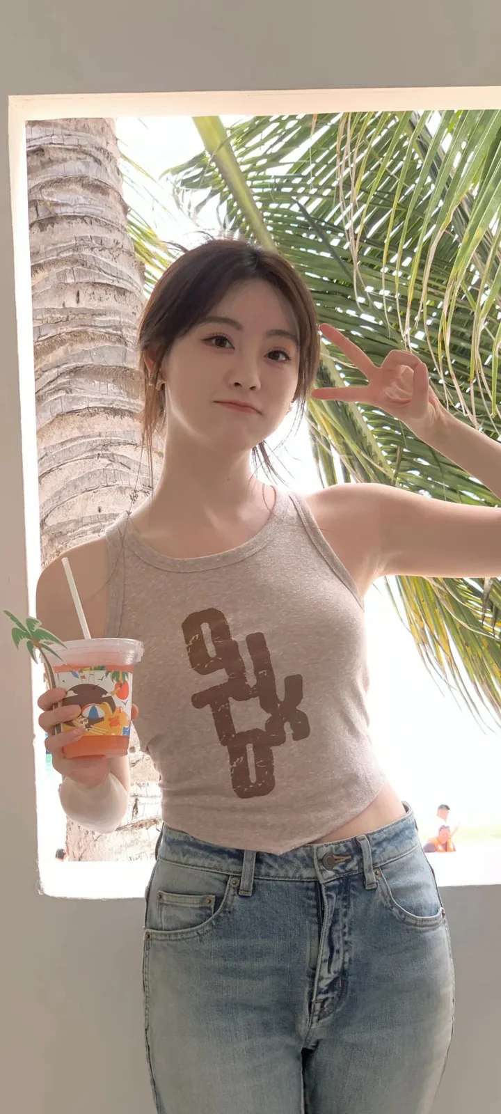 一名女子站在棕榈树旁，手持饮料，沐浴在明亮的户外光线中。