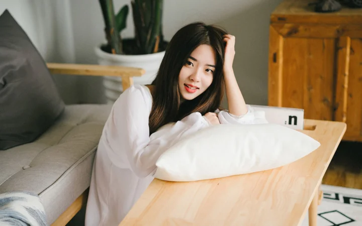 흰 셔츠를 입은 성인 여성이 실내에서 소파 옆 테이블에 기대고 있습니다.