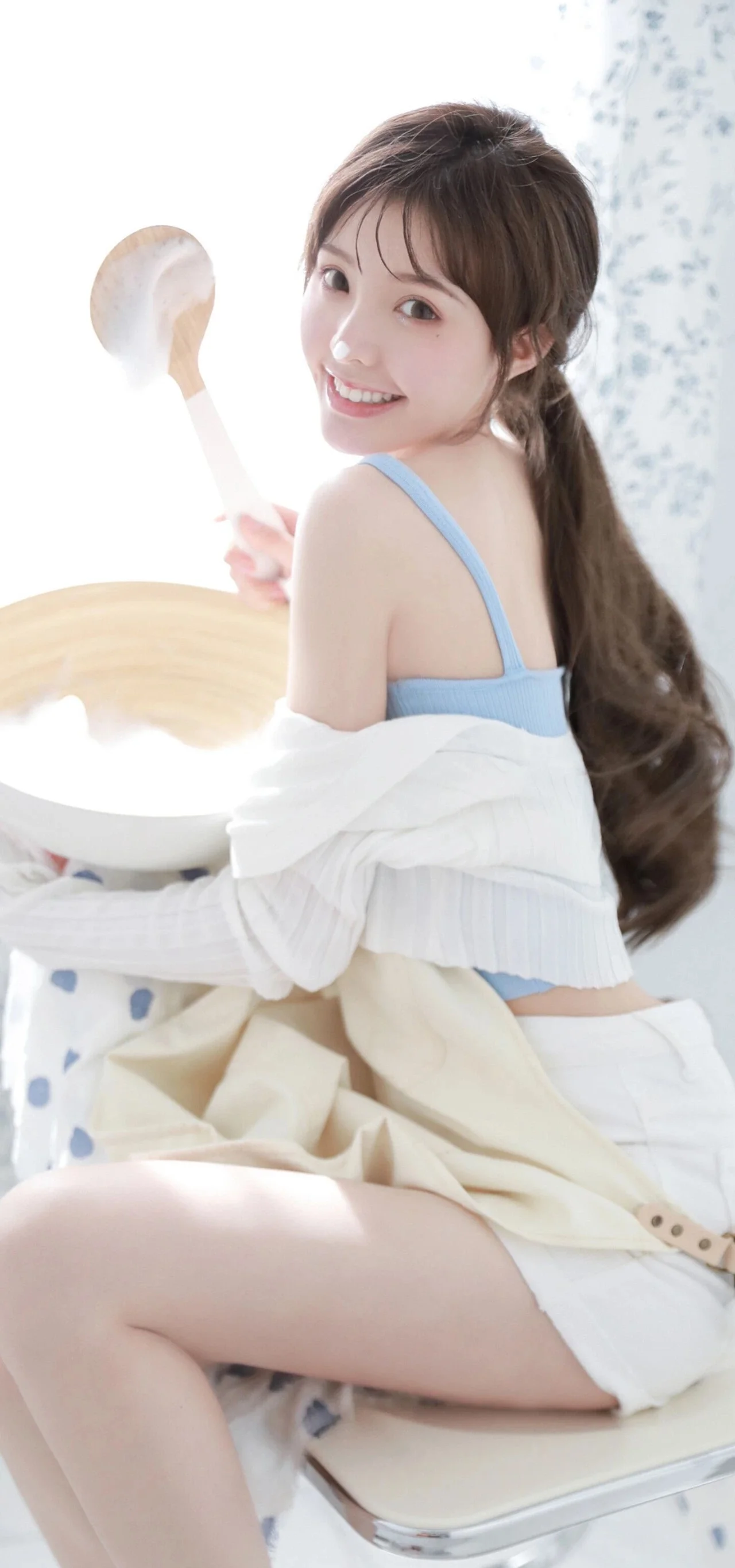 一名長髮女性回頭微笑，手持湯匙坐在室內淺色背景前。