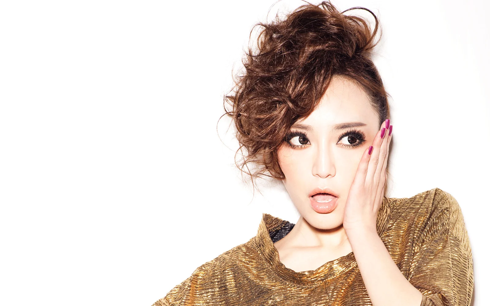 一名妆容精致、卷发造型的成年女性身穿金色上衣，在白色背景前摆姿势。