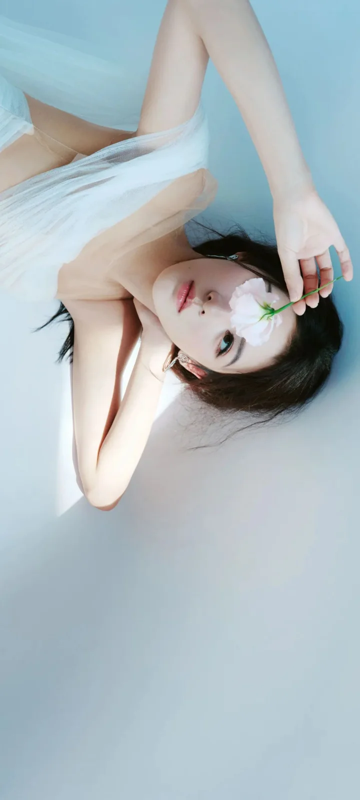 淡い青色の背景に横たわる成人女性が、白い花を手にしている。