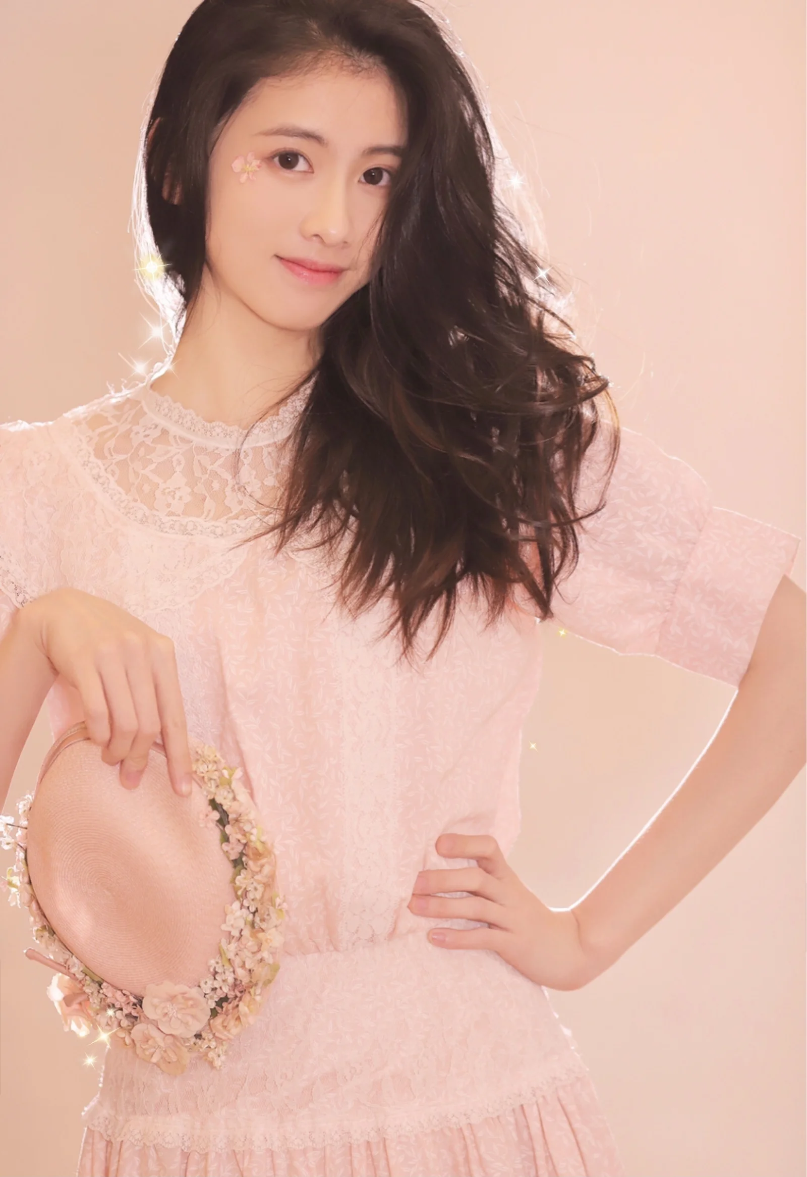 長い髪の女性が淡いピンクのドレスを着て、花飾りの帽子を持ちながら柔らかなピンク色の背景でポーズを取っている。