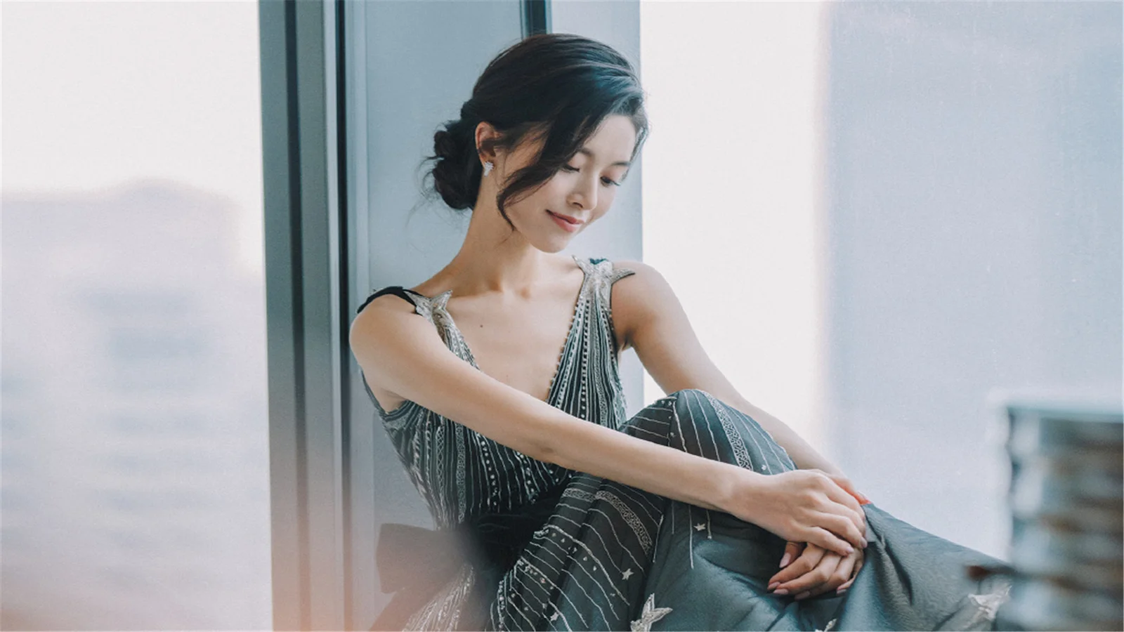 一名成年女子坐在窗边，低头看向一侧，柔和的自然光照亮了她和室内空间。