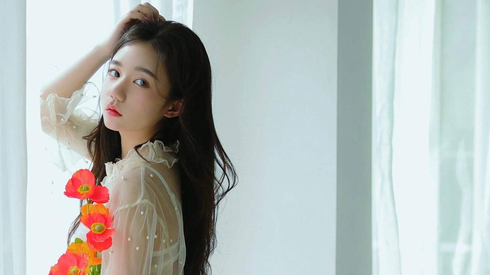 一位长发女子站在明亮窗边，手扶头发，身旁映衬着红色花朵。