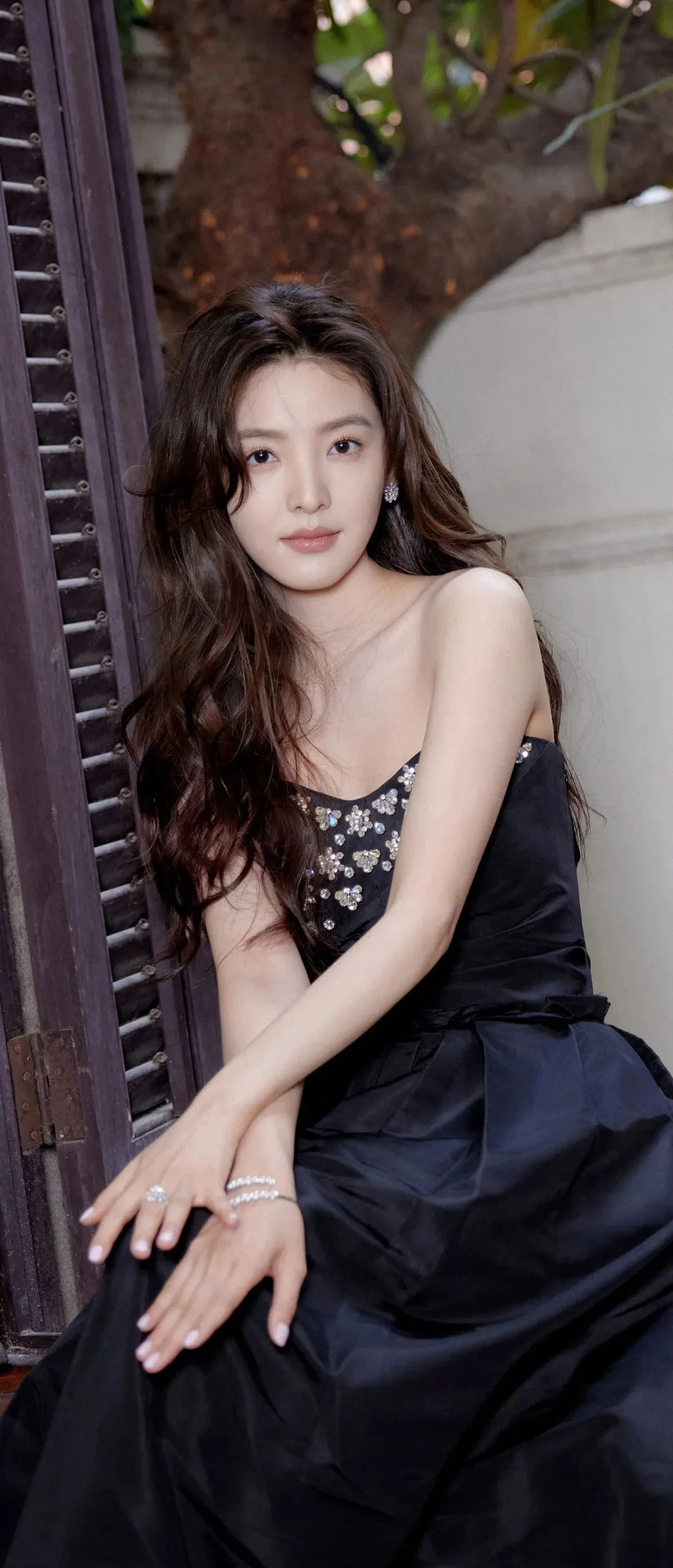 검은 드레스를 입은 성인 여성이 야외의 나무와 건물 옆에서 포즈를 취하고 있다.