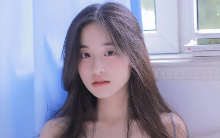 긴 머리의 여성이 창가에서 카메라를 바라보며 부드러운 파란색과 분홍색 조명을 받고 있습니다.